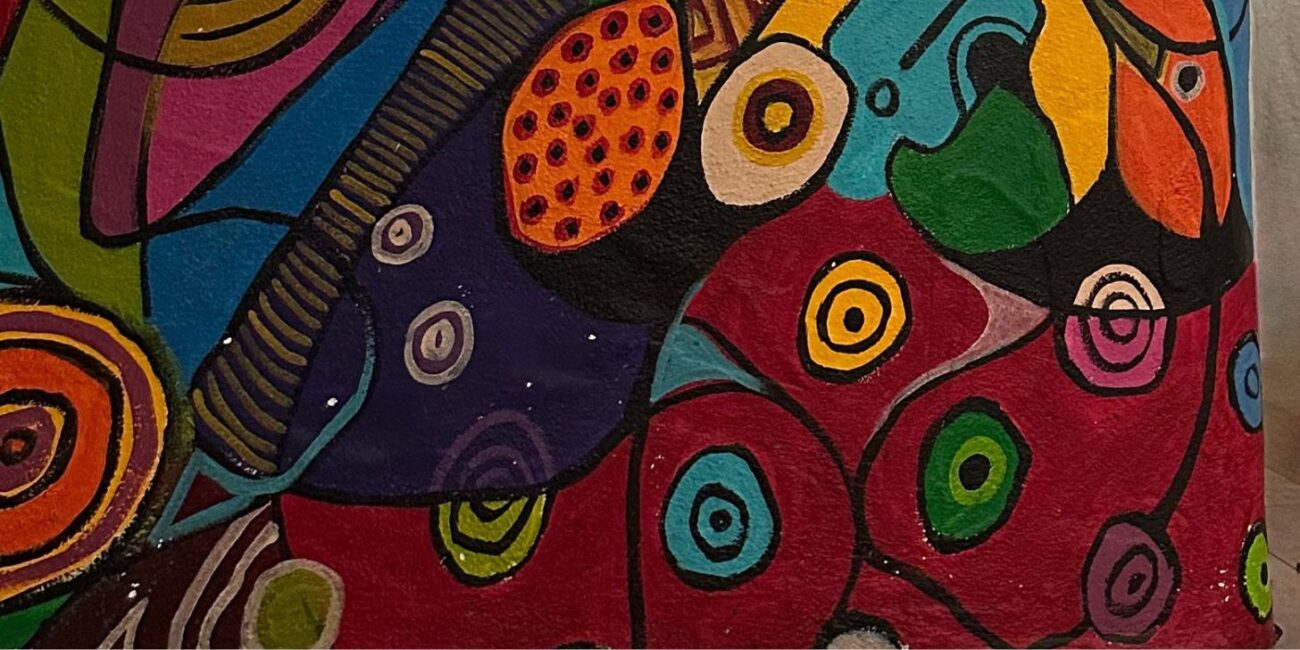 Photographie en gros plan d'une surface peinte aux couleurs vives (rouge, violet, turquoise, jaune) présentant une multitude de cercles concentriques évoquant des cellules ou des yeux, ainsi que des zones pointillées rappelant des écorces de fruits ou des textures animales.