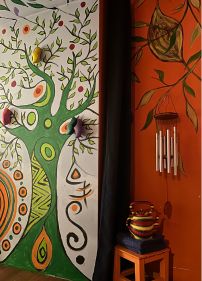Vue d'un angle du sanctuaire montrant un grand arbre de vie peint sur un mur blanc avec un tronc vert et des branches s'étendant vers le plafond, jouxtant un mur orange vif orné de motifs végétaux et d'un carillon à vent suspendu au-dessus d'une poterie colorée.