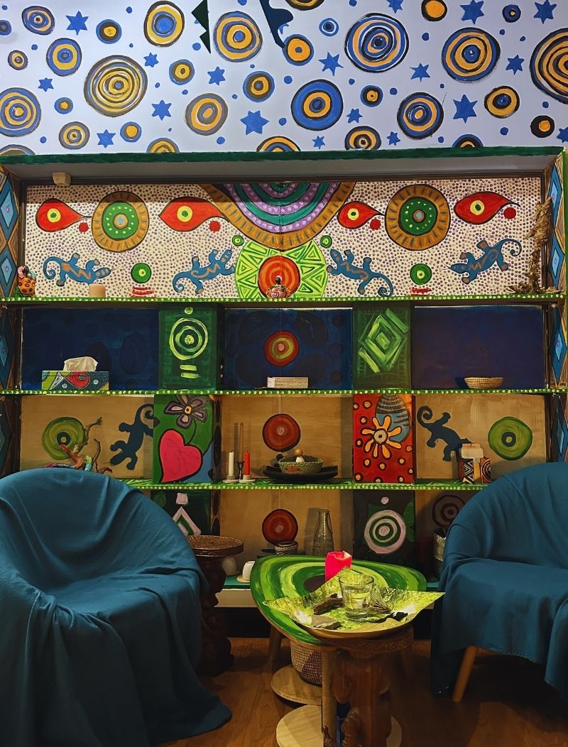 Vue d'un espace de soin intérieur décoré par Marguerite Lalèyê, comprenant une bibliothèque peinte de motifs de lézards, de cœurs et de cercles vibrants, surmontée d'une fresque murale de cercles et d'étoiles bleues sur fond clair, avec deux fauteuils bleus invitant à la confidence