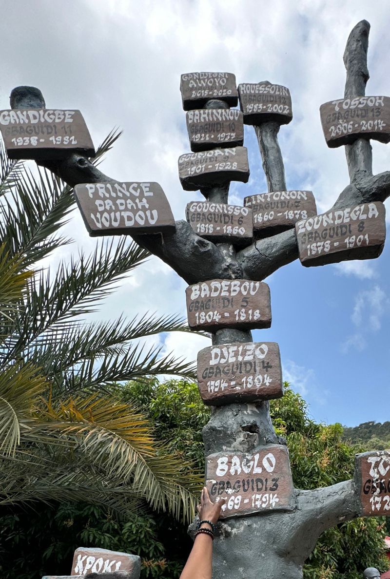Photographie en contre-plongée d'un monument en forme d'arbre généalogique sculpté sous un ciel nuageux. On aperçoit uniquement la main de Marguerite Lalèyê, ornée d'un bracelet de perles sombres, venant toucher la plaque dédiée au Roi Gbaguidi Baglo (1769-1794). De nombreuses autres plaques portant les noms des rois successifs sont fixées sur les branches de la structure.