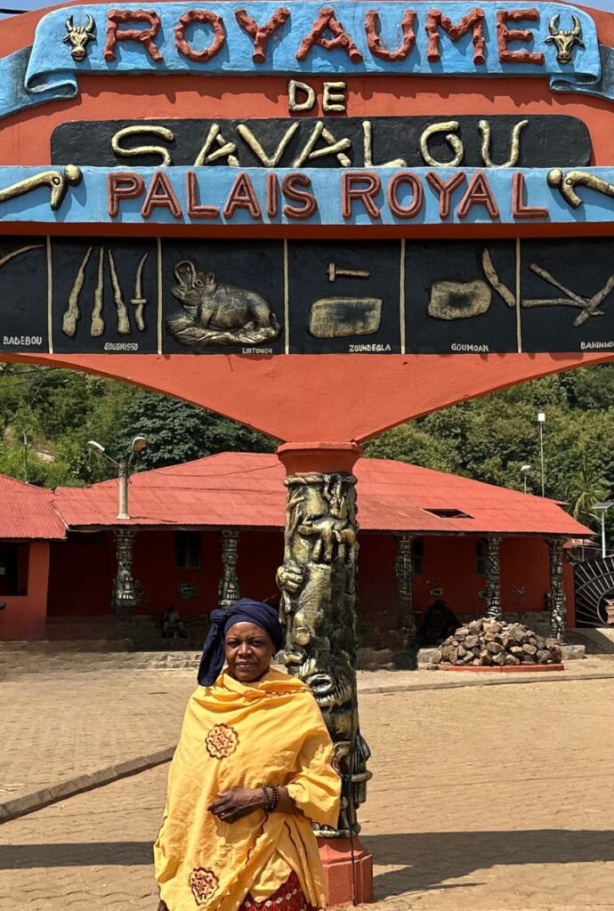 Photographie de Marguerite Lalèyê posant devant une entrée monumentale orange et bleue du Palais Royal de Savalou. Elle porte une tenue traditionnelle avec un grand drapé jaune orné d'un motif circulaire et un turban bleu foncé. En arrière-plan, on aperçoit des bâtiments à toit rouge et une végétation luxuriante sous un ciel clair.
