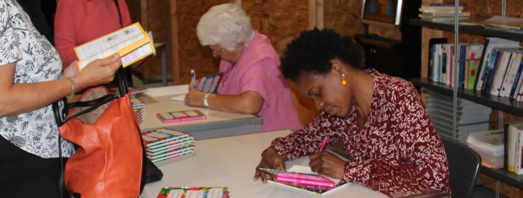 Photographie de Marguerite Lalèyê et Rosette Poletti assises à une table lors d'une séance de dédicaces. Marguerite, au premier plan à droite, porte un chemisier à motifs bordeaux et écrit dans un exemplaire du "Petit carnet de pensées". Rosette Poletti, à ses côtés, dédicace également un ouvrage. Une lectrice se tient debout à gauche, feuilletant le livre.